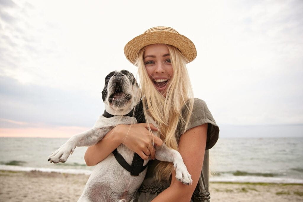 Funny shot of beautiful young woman with long blonde hair wearing casual clothes, walking along a dog friendly off leash beach on overcast day with her dog, looking at camera joyfully and smiling widely