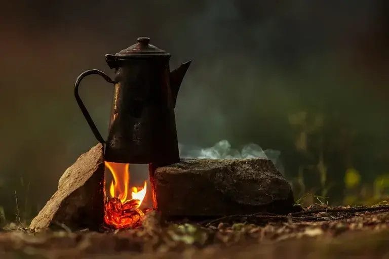 Coffee is brewed on a small fire in a rustic red camping kettle.