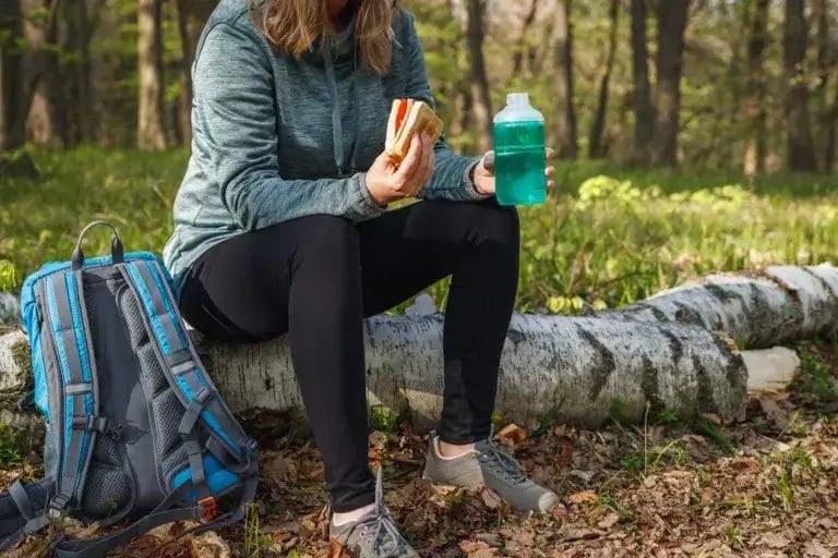 Woman eating sandwich and drinking water outdoors.