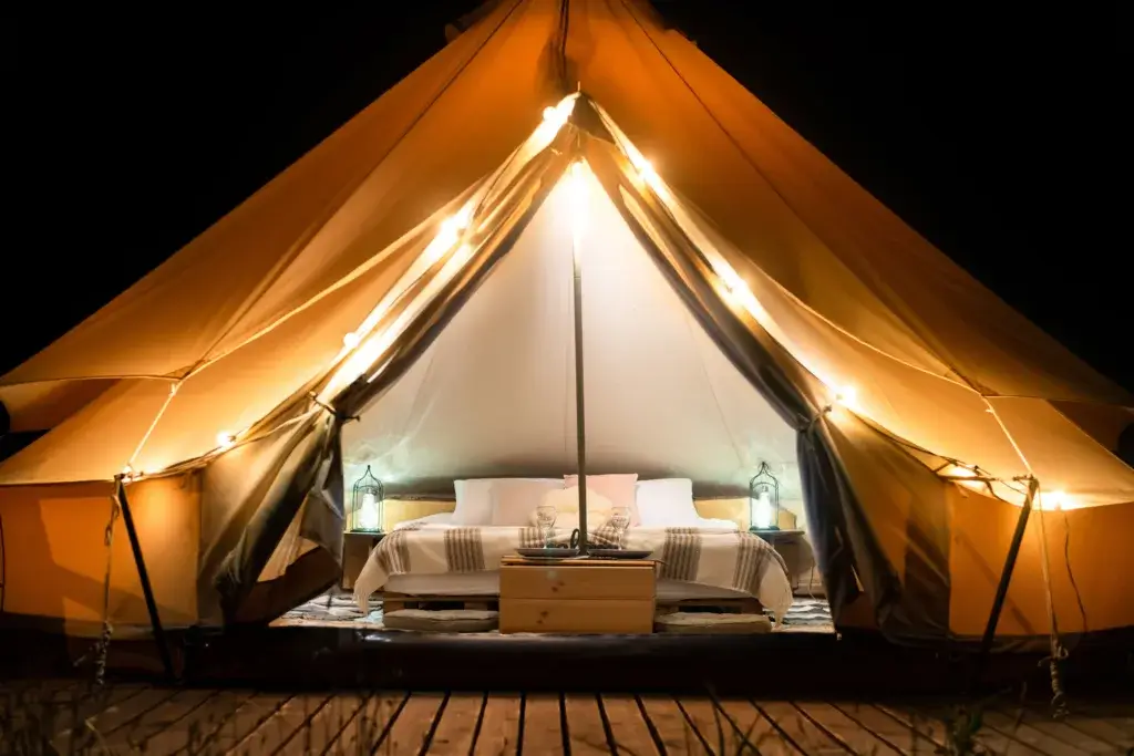 Tent interior and exterior with lamps, bed and furniture glamping at night.