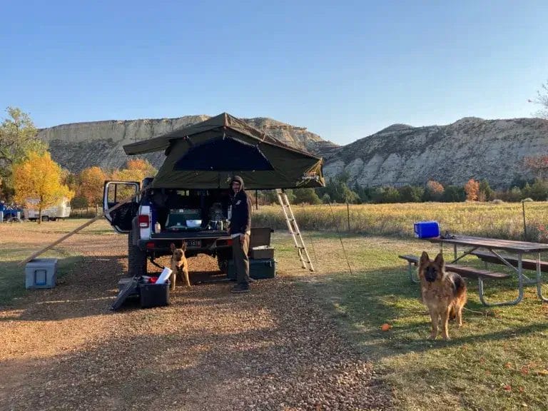 A man with two German Shepherd dogs standing by a Jeep Gladiator with a rooftop tent in a campground near Badlands National Park.