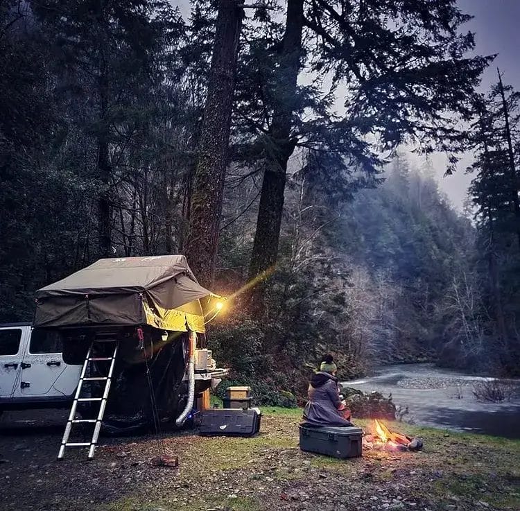 A woman sitting next to a campfire next to the river with a rooftop tent on a Jeep Gladiator in the background.