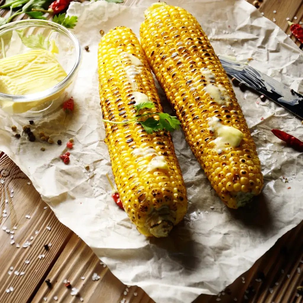 Top view of kitchen table with grilled sweet corn cob under melting butter and greens on baking paper.