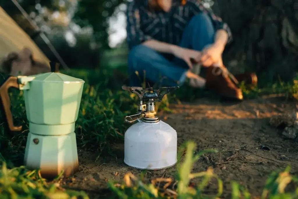 A portable propane burner and geyser coffee maker are in the foreground for making coffee for the tourists. Tourism and recreation in the forest in summer with a tent. Tourist in the background.