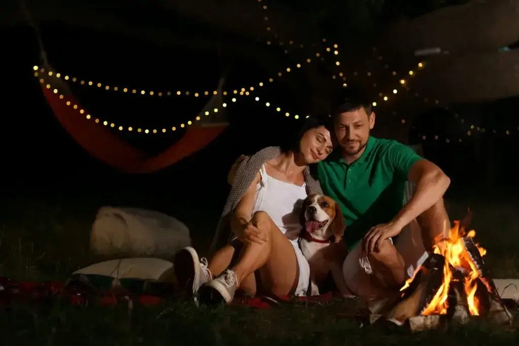 Happy couple with dog near fire on summer evening.