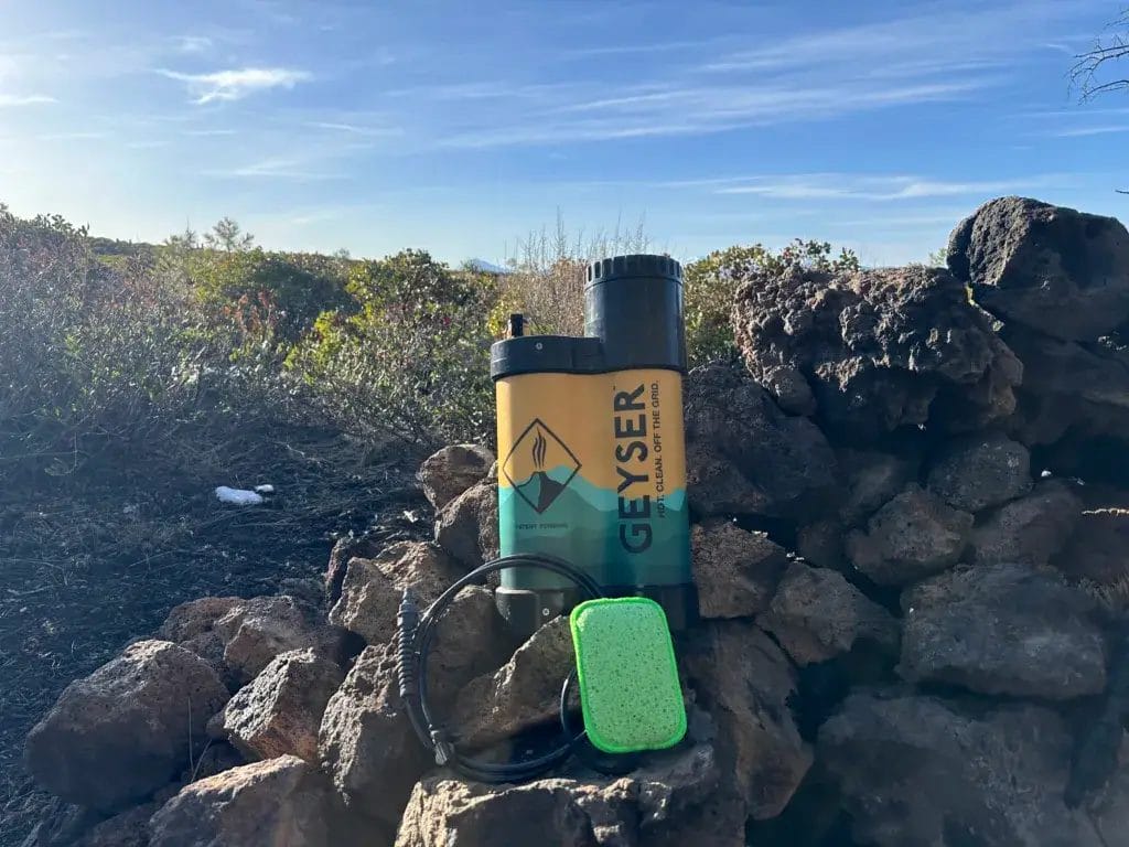 Geyser camping shower with hose and sponge sitting on a campfire ring in the wilderness.