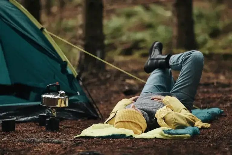 Woman solo camping in hiking gear lying on her sleeping bag at her campsite after a night camping in the woods.