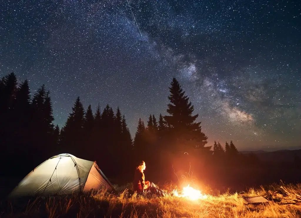 Rear view of woman hiker sitting by bright burning campfire near tent alone, thinking and enjoying beautiful camping night under dark sky full of stars and bright Milky Way, warm summer 