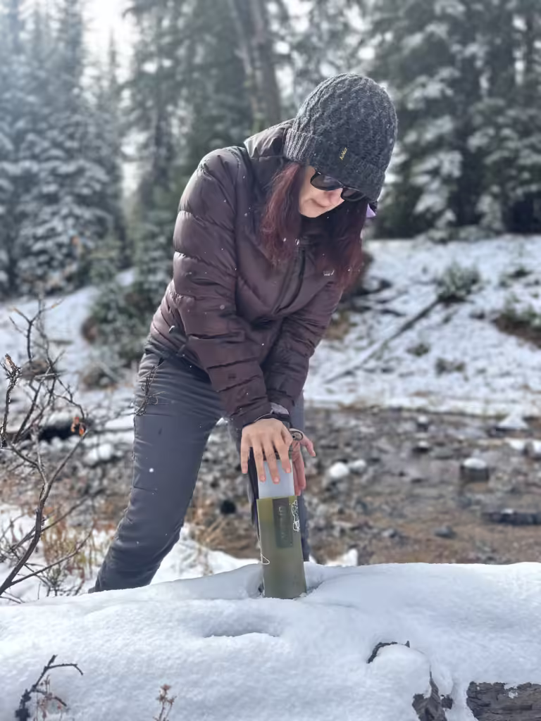 Woman filtering water with a GRAYL purifier in a snowy stream.
