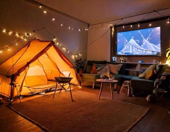 A cozy living room with a tent pitched in the center, surrounded by string lights and camping equpment and a movie projector.