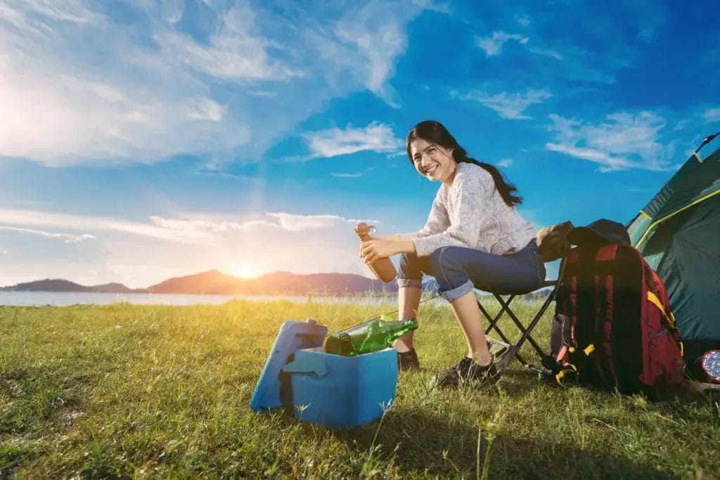 Smiling young woman sitting on a chair in a grassy field by a lake solo camping.