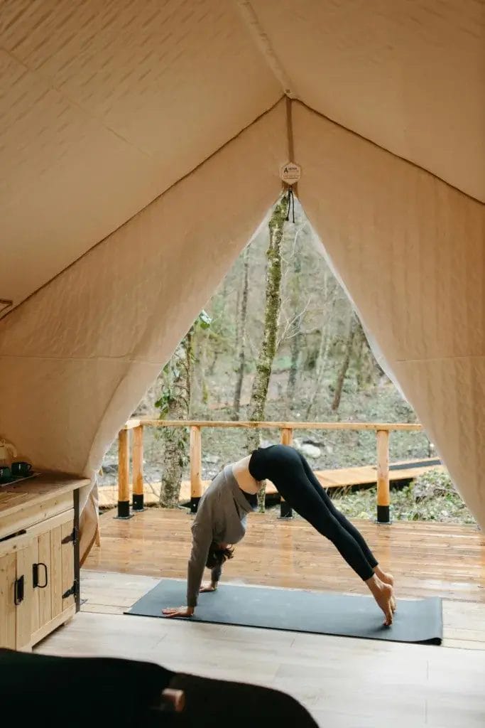 A woman practicing yoga on a mat inside a cozy tent cabin with a forest view.