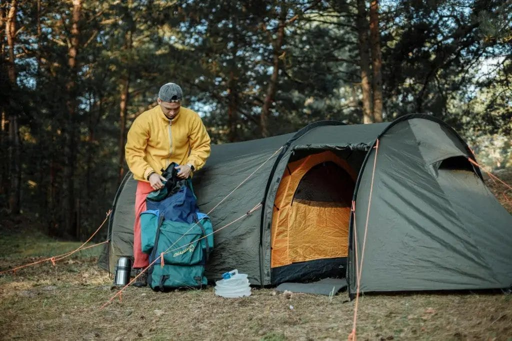 A young man organizing camping gear beside a tent in a scenic forest.