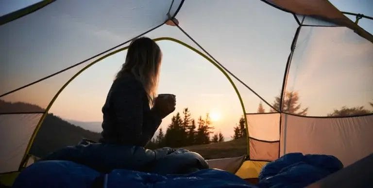 Woman in tent with a cup of coffee in her hand watching the sun rise.