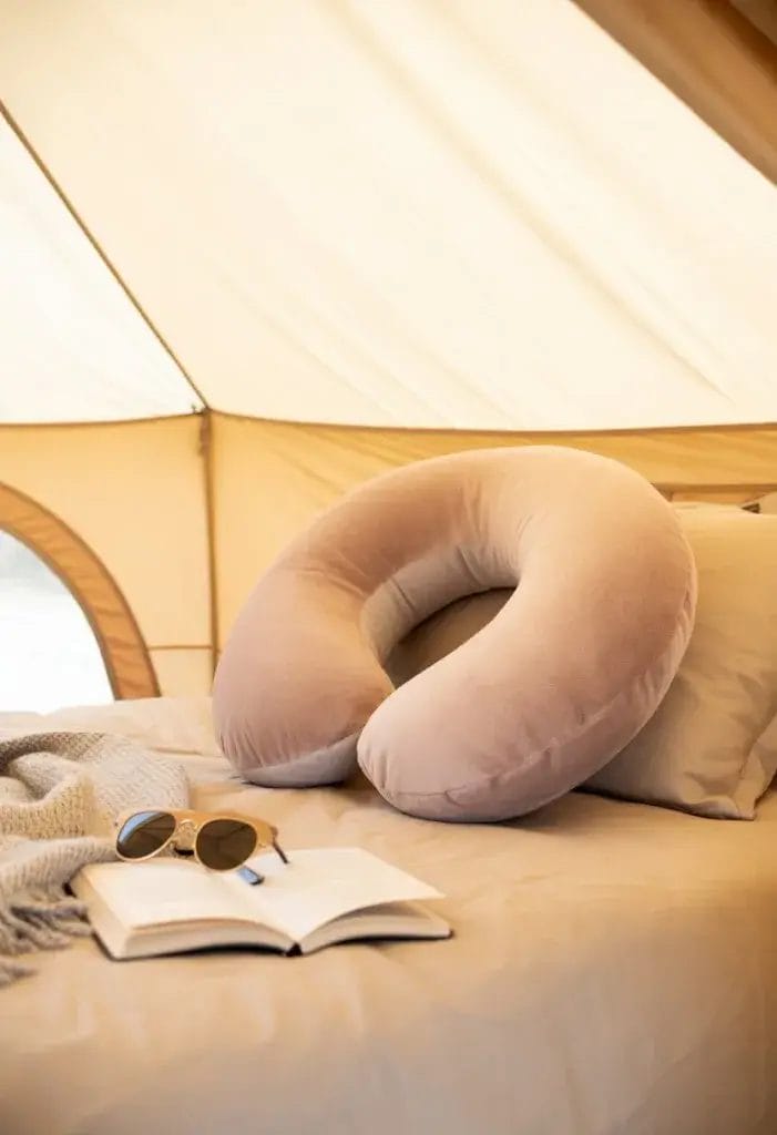 Inside a cozy glamping tent, a soft neck pillow—plush and inviting, perhaps in a pastel or neutral color—rests on a comfy bed or camping chair. The pillow looks supportive and perfect for napping, suggesting comfort and relaxation. Nearby, there might be a lightweight blanket, a book, or a pair of sunglasses, enhancing the restful vibe. Soft, natural light filters through the tent, and the overall scene feels warm, homey, and peaceful—showing how a simple neck pillow can make naps in the tent feel just like home.