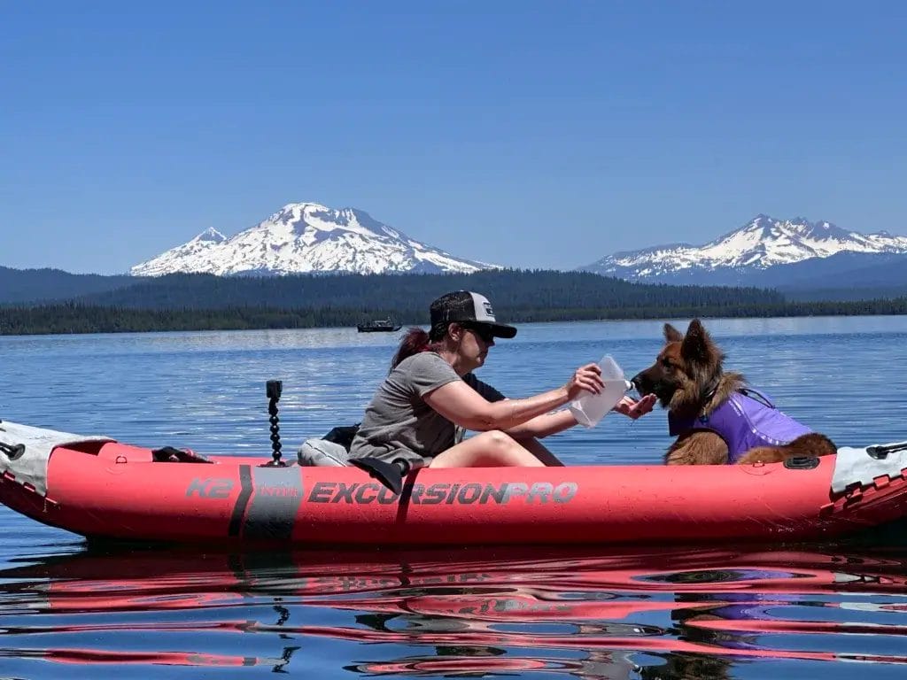 Woman giving water to a German Shepherd dog in a kayak with a scenic mountain view in the background.