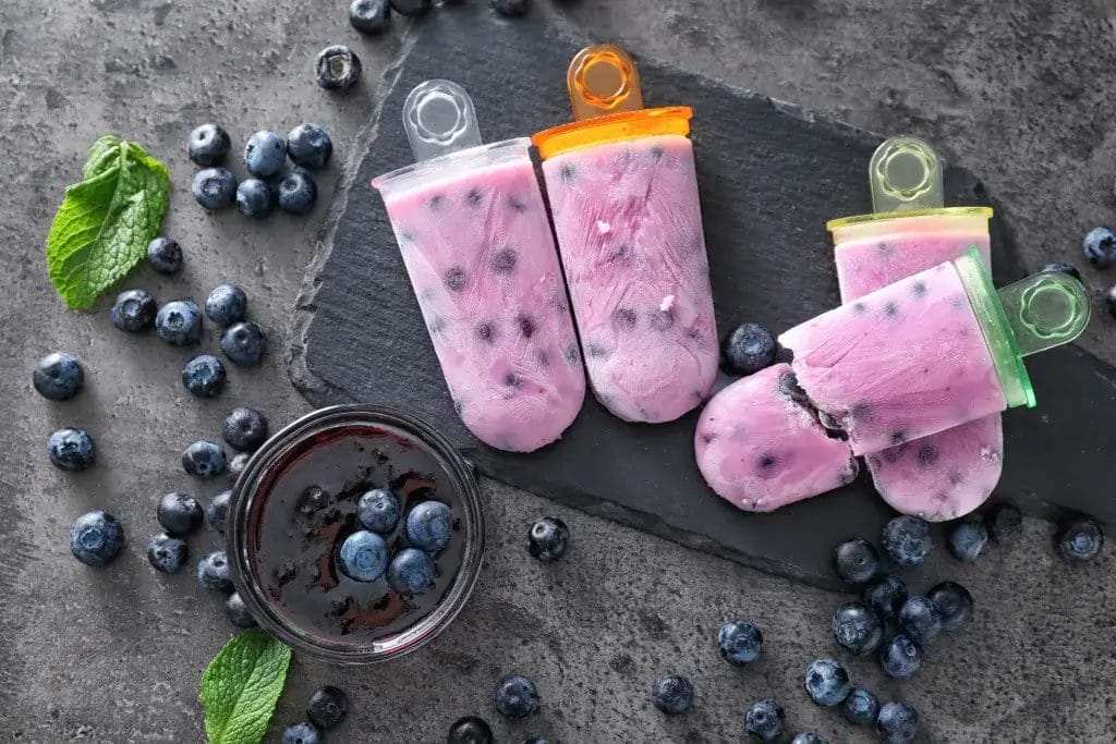 Yogurt blueberry popsicles on a table with fresh blueberries lying around.