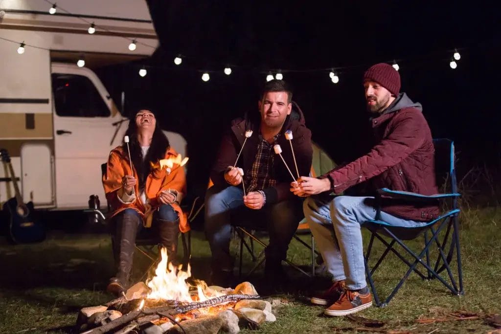 Three friends are sitting around a campfire roasting marshmallows with an RV motorhome in the background.