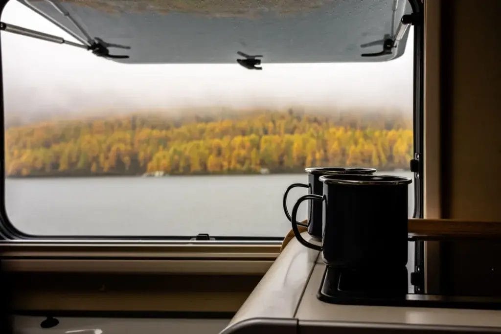 Two mugs sitting on a table in a RV with autumn colors outside the window.