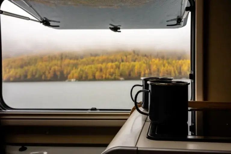 Two mugs sitting on a table in a RV with autumn colors outside the window.