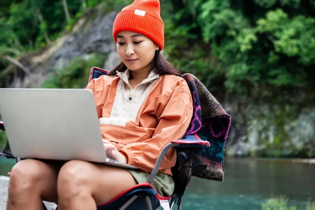 Woman sitting in a camp chair next to a creek working remotely on a laptop.