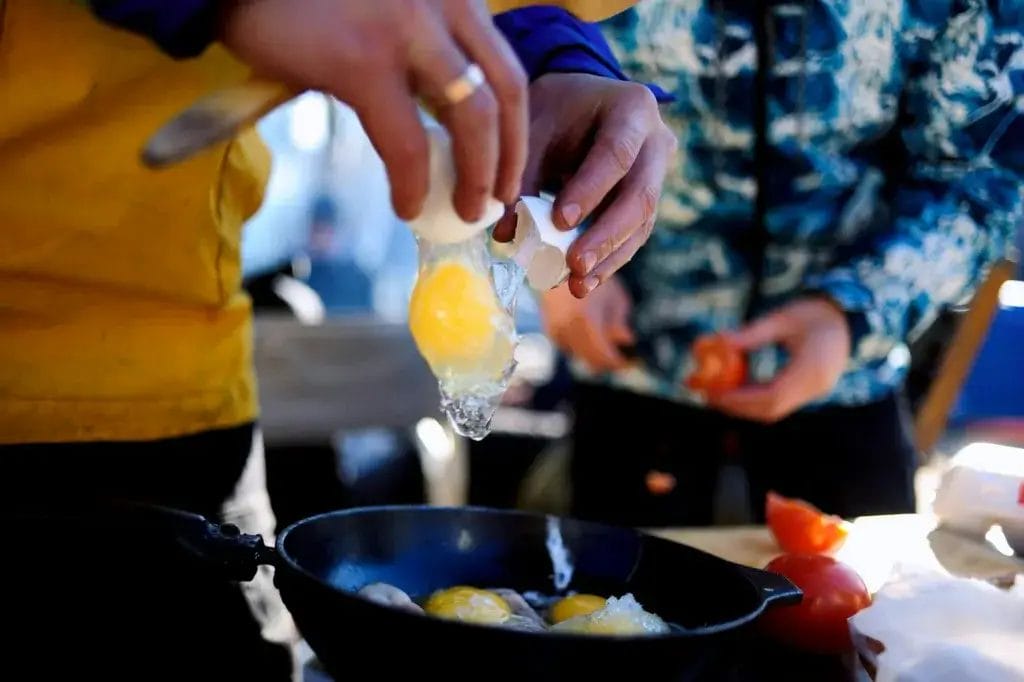 A person cooking eggs in a frying pan outdoors, focusing on ingredients.
