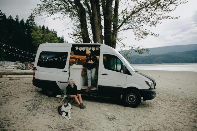 A couple with their dog relax by a camper van on a serene beach, embracing outdoor lifestyle.