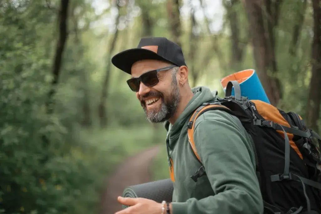A man smiling wearing sunglasses and a backpack on a hiking trail.