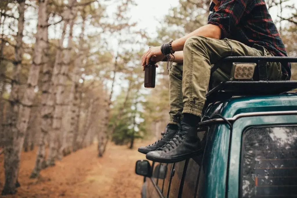 A man dressed in outdoor gear is sitting on a car roof in the woods.